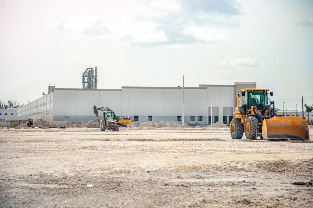Construction site with yellow excavator and loader moving earth in front of large industrial building
