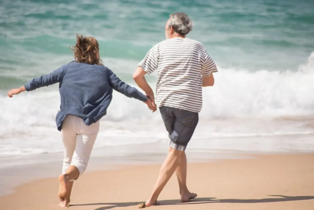 Older couple running hand-in-hand on a sandy beach with ocean waves in the background