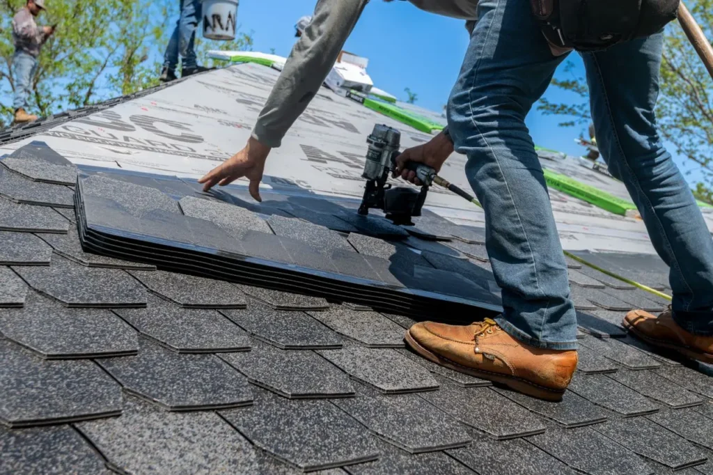 Roofer installing black shingles on residential roof with nail gun under clear blue sky