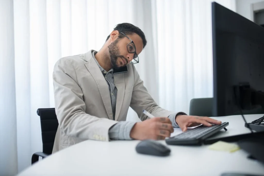 Professional man in beige blazer multitasking on phone call while writing notes and using computer at desk