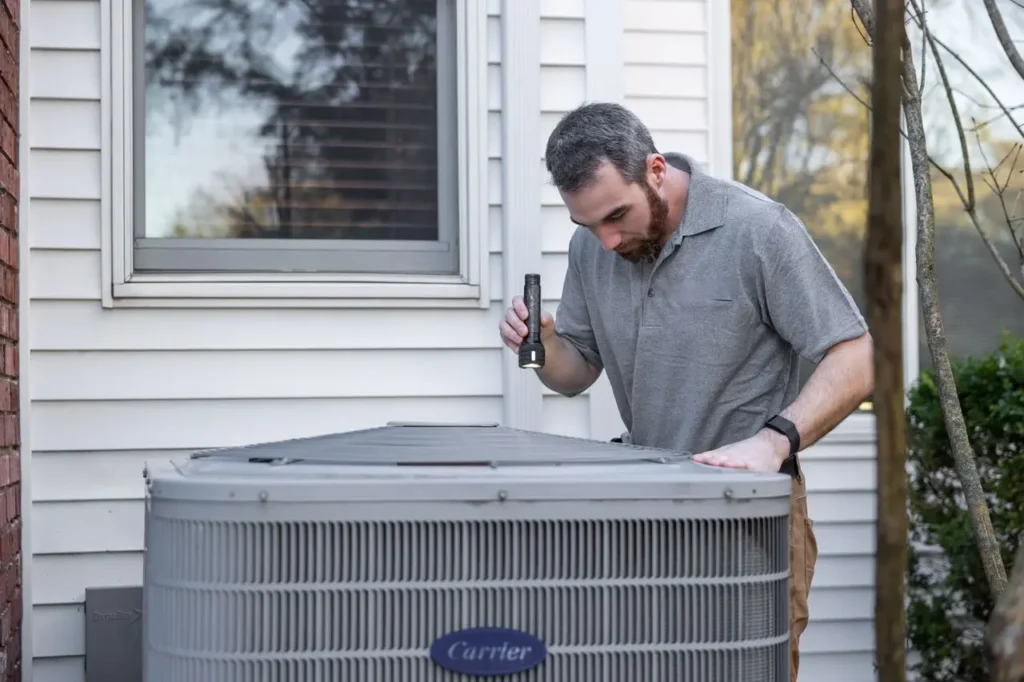 Technician inspecting a Carrier air conditioning unit outside a home while holding a flashlight