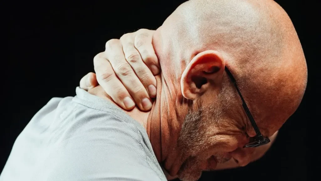 Bald man with glasses holding his neck and shoulder in pain against black background