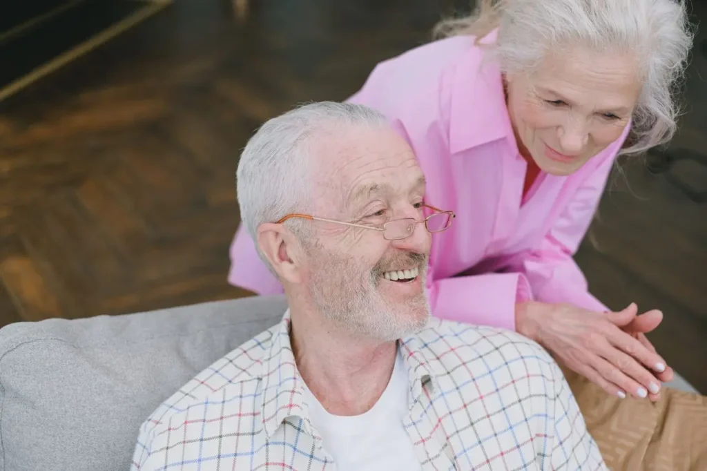 Senior couple smiling together, woman in pink placing caring hand on man's shoulder as he sits on couch